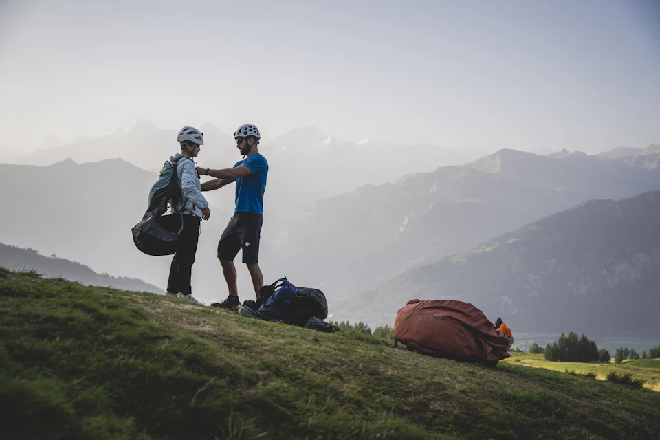 Two people in helmets prepare gear on a grassy hilltop, with mountainous scenery in the background.
