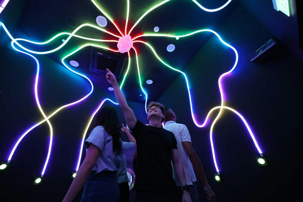 A group of people standing under colorful neon lights forming branching patterns on the ceiling in a dark room.