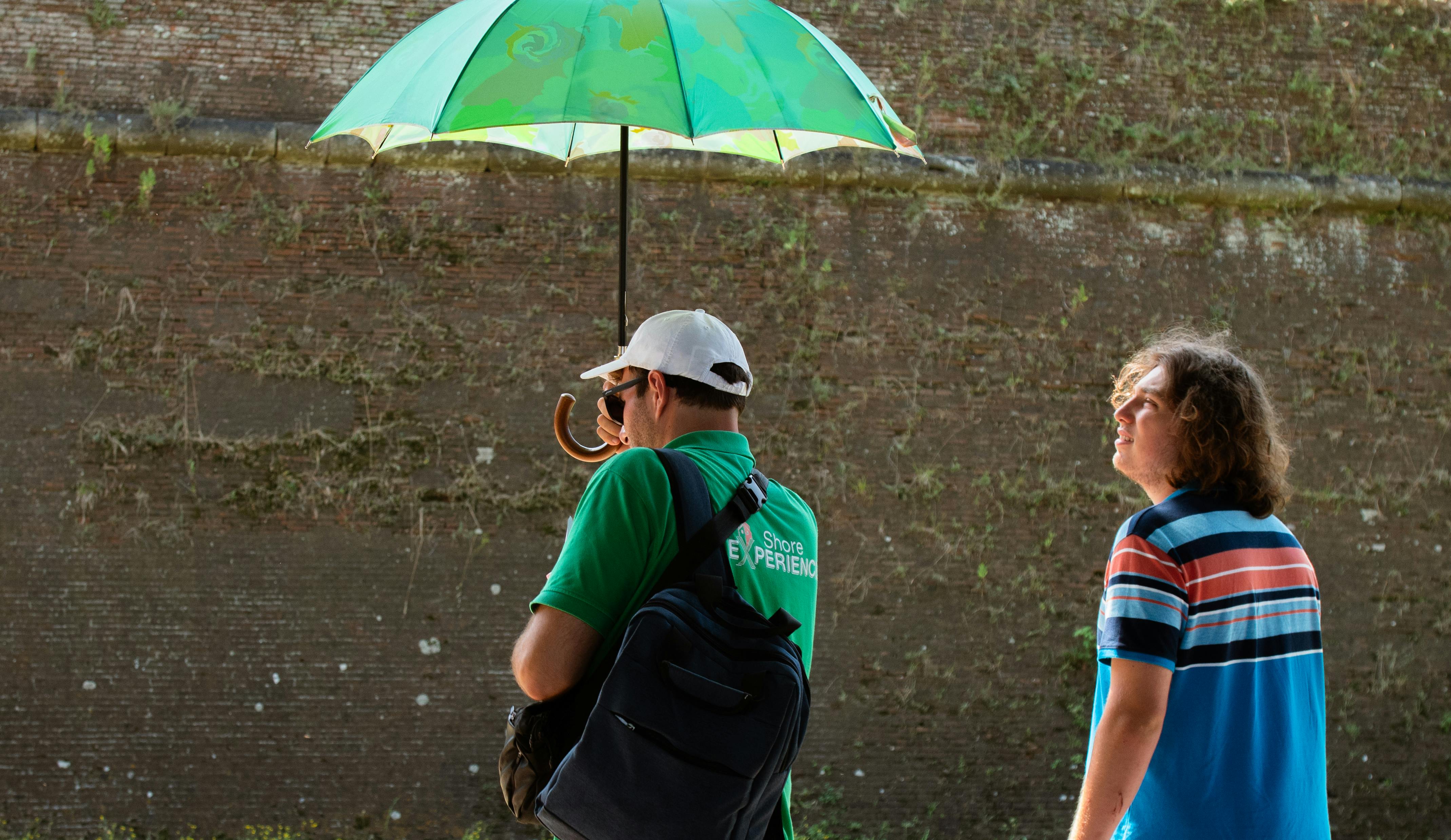 Two people stand outdoors near a moss-covered wall. One holds a green umbrella, and the other, wearing a striped shirt, looks up.