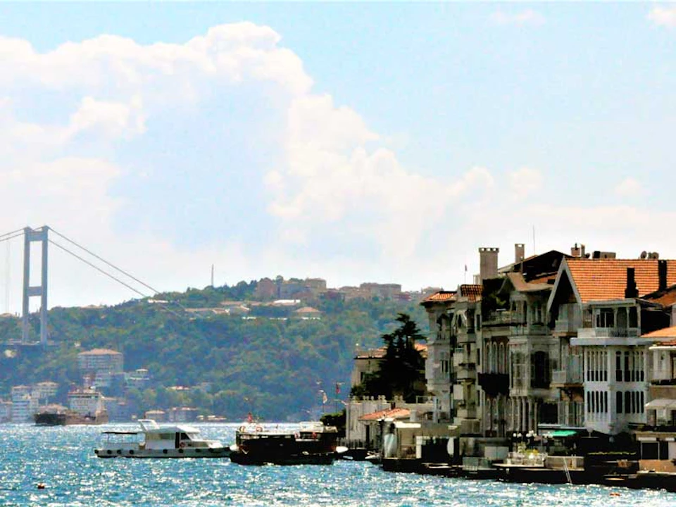 Waterside view of a coastline with buildings, boats anchored, and hills in the distance under a cloudy sky.