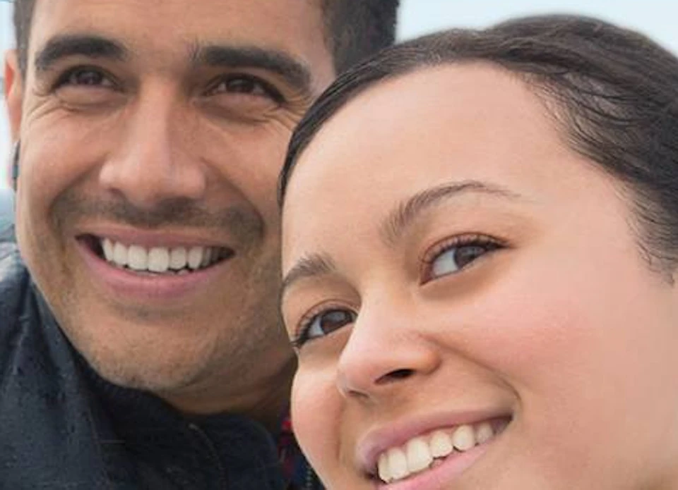 Two people smile and pose on a boat with the Golden Gate Bridge in the background. One points while the other holds a phone.
