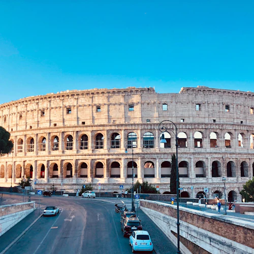 Colosseum i Rom badade i solljus, med en klarblå himmel, bilar parkerade i närheten och några personer som gick längs gatorna.
