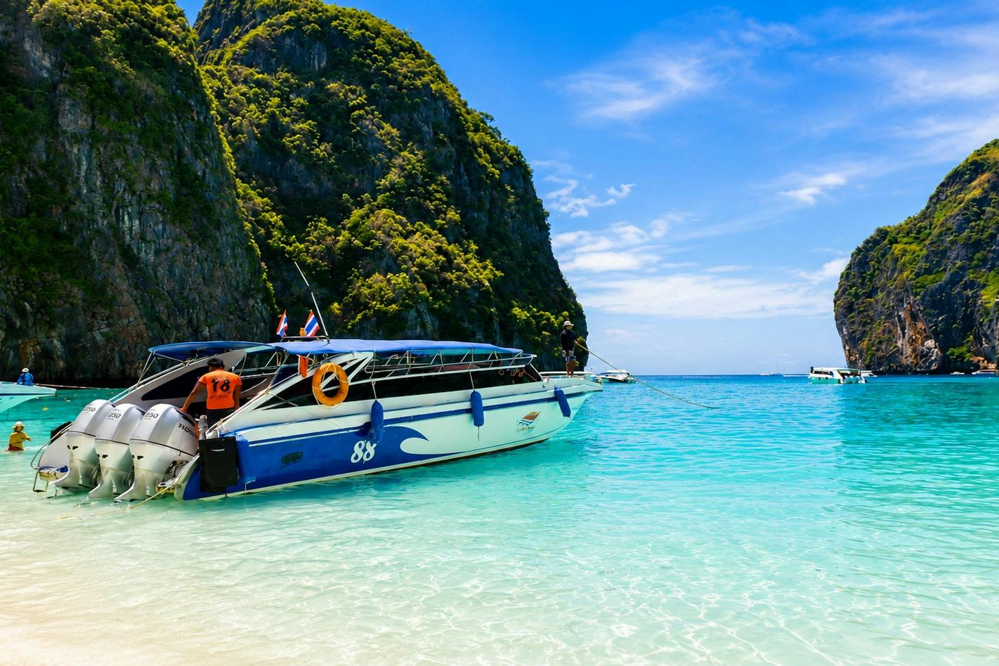 A speedboat docked at a tropical beach with clear turquoise water, surrounded by lush green cliffs under a blue sky.
