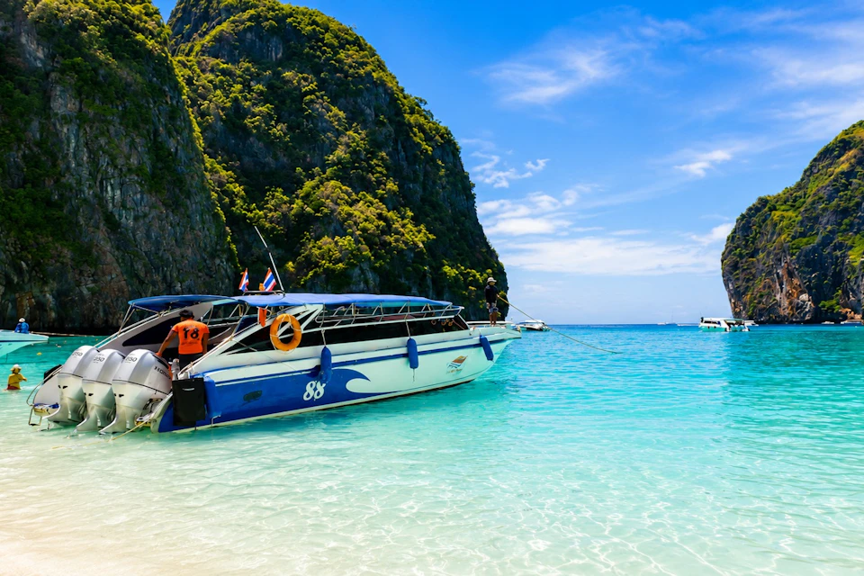 A speedboat docked at a tropical beach with clear turquoise water, surrounded by lush green cliffs under a blue sky.
