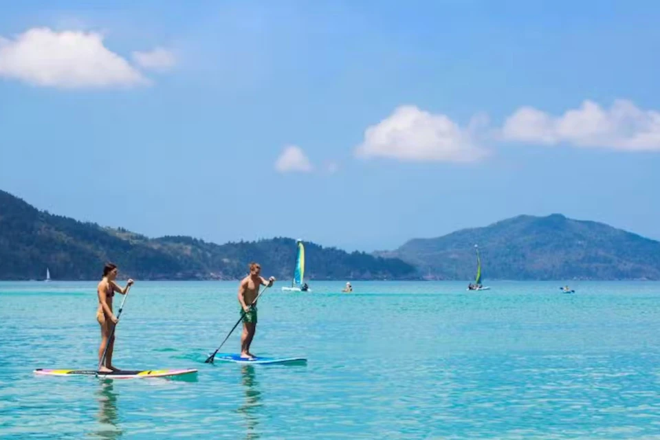 Two people paddleboarding on clear blue water with distant sailboats and green hills under a sunny sky.