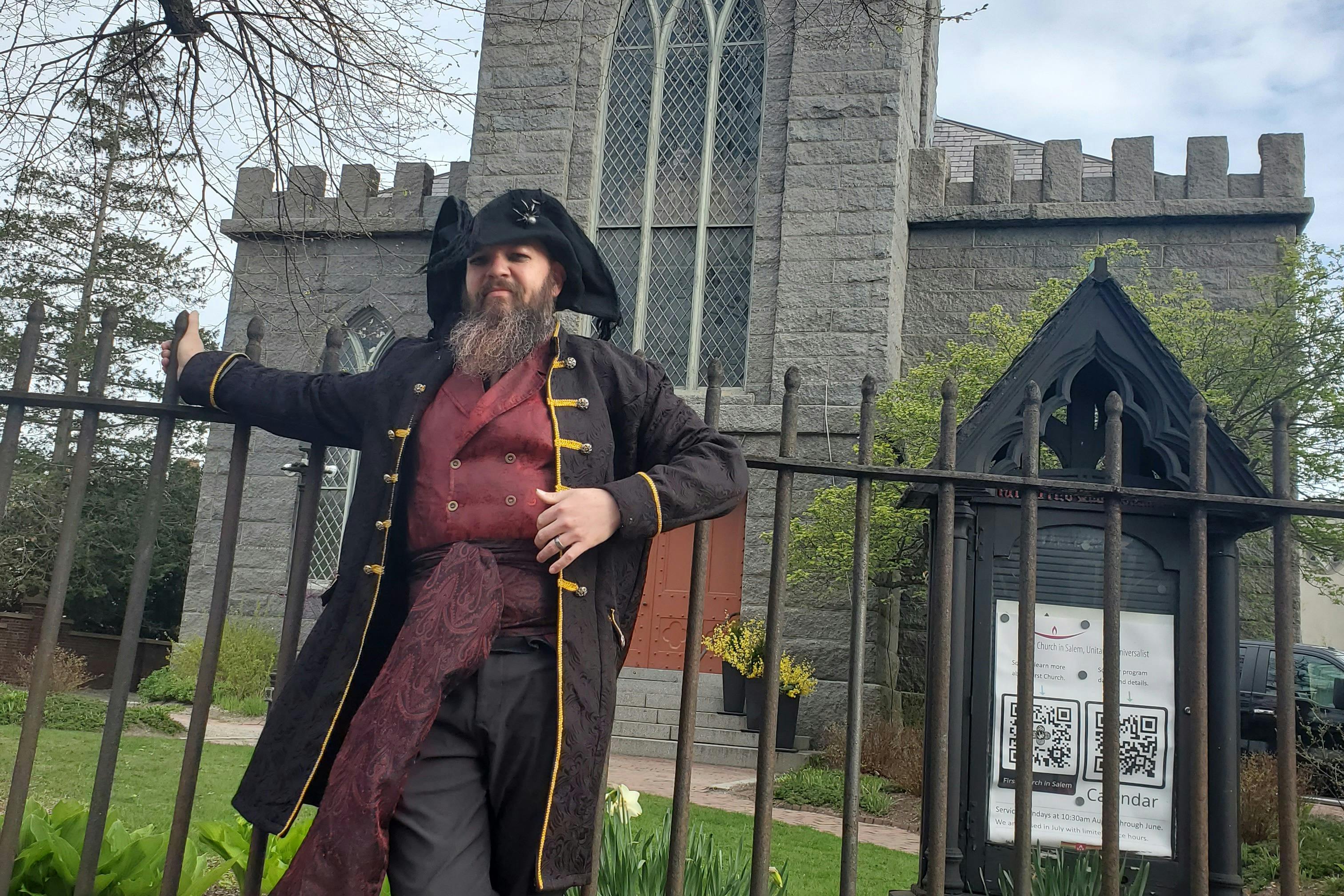 Ένας άντρας με καπέλο και παλτό από τρικόρνο στέκεται δίπλα σε έναν φράχτη από σφυρήλατο σίδερο μπροστά από ένα πέτρινο κτίριο με τοξωτά παράθυρα.