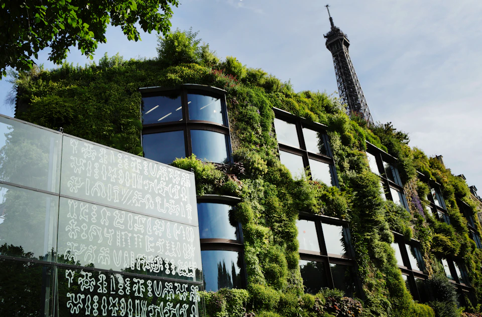 A building with a green wall of plants, large windows, and an adjacent panel with symbols. The Eiffel Tower is visible in the background.