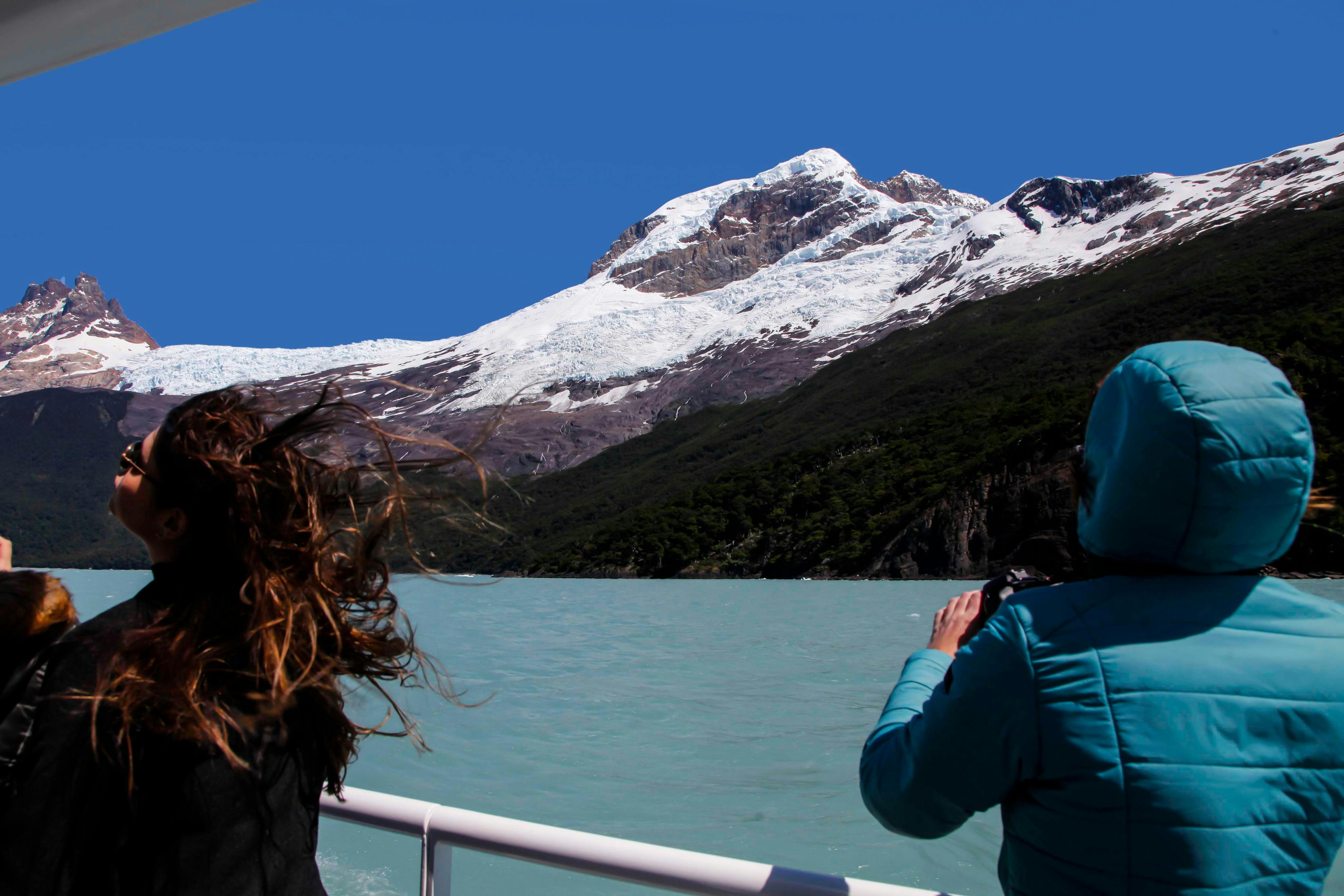 Two people on a boat admire a glacier-covered mountain surrounded by greenery under a clear blue sky.