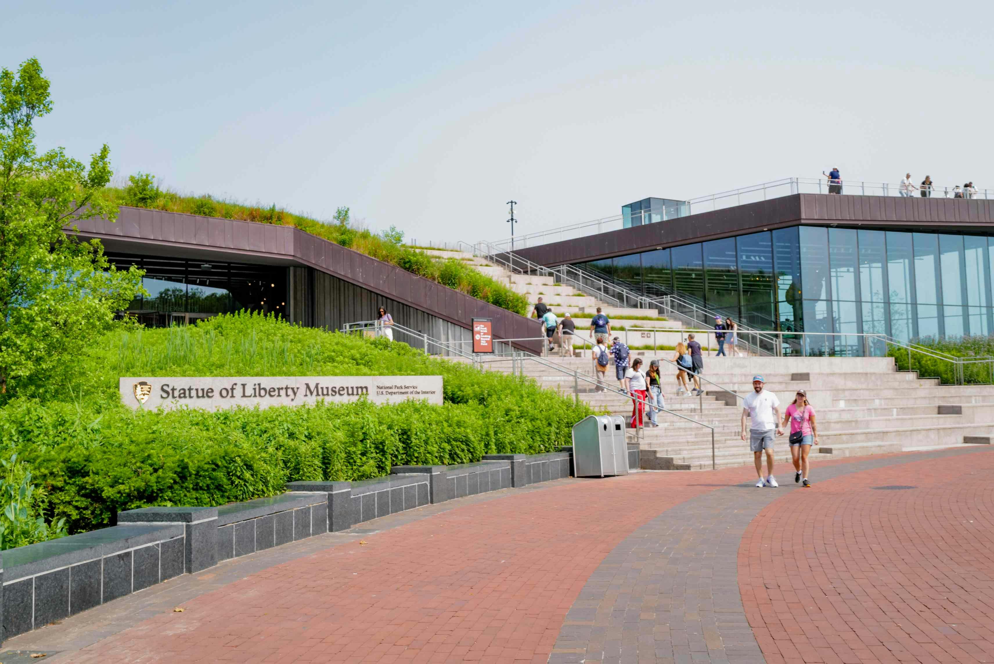 Besøgende, der går i nærheden af indgangen til Frihedsgudinden, er omgivet af grønne landskaber og brolagte stier.