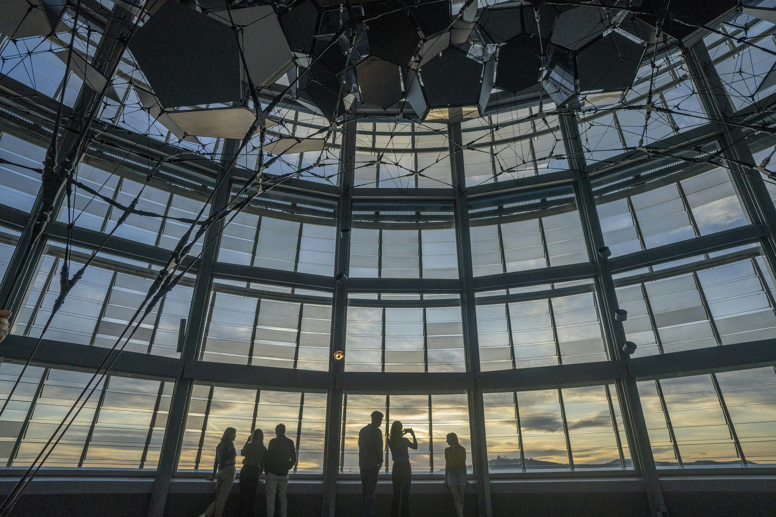Silhouettes of people stand inside a large glass-domed structure, with a sunset visible through the windows.