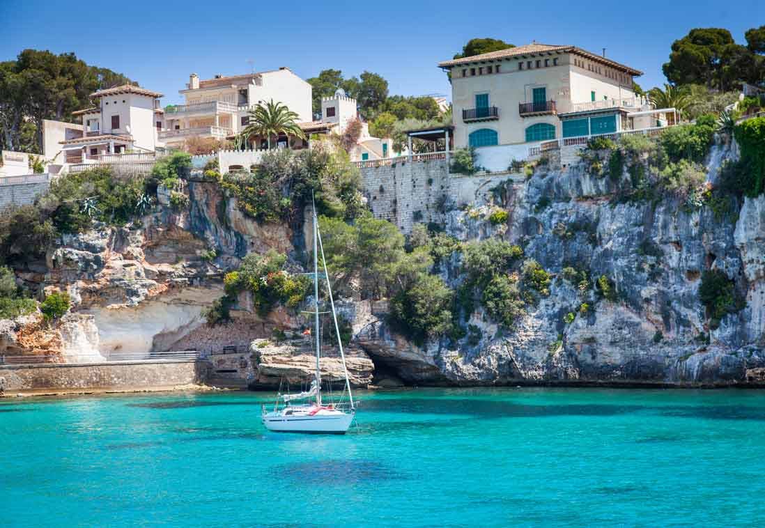 一艘白色帆船漂浮在清澈的绿松石水面上，靠近岩石悬崖，上面有房屋和绿色植物。