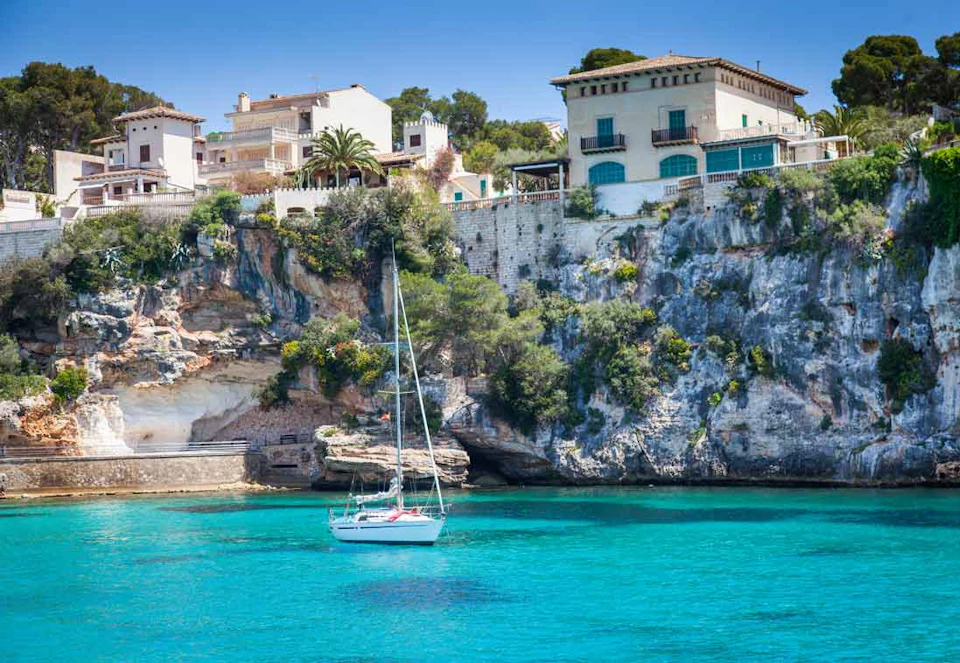 A white sailboat floats on clear turquoise water near a rocky cliff with houses and greenery above.