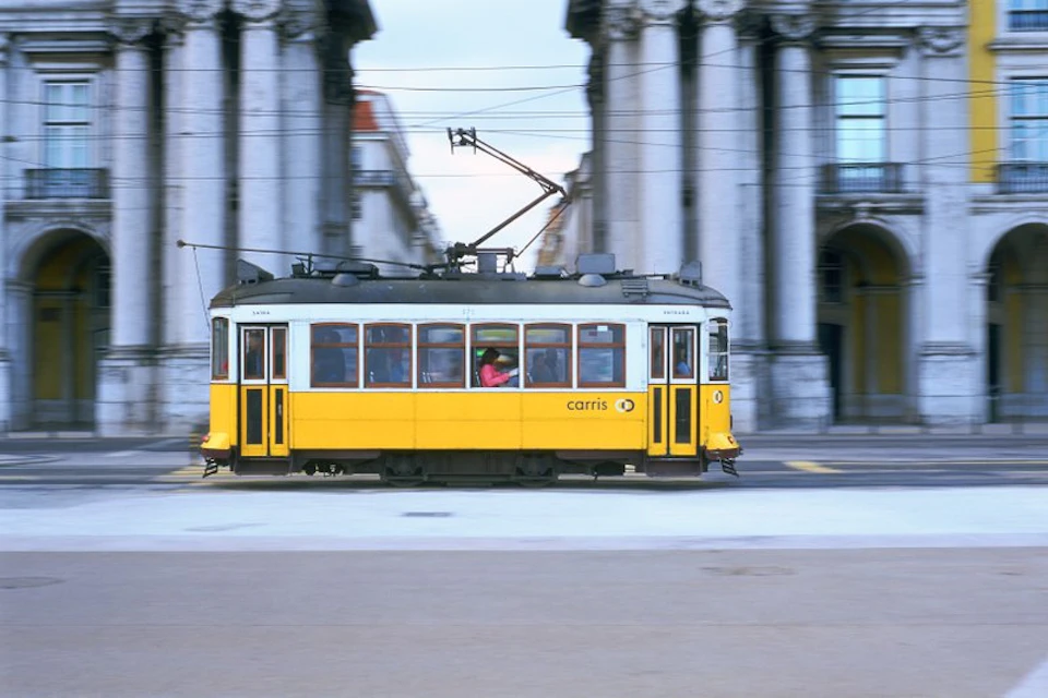 A yellow tram passes by gray, columned buildings, with a person in pink visible through the windows.