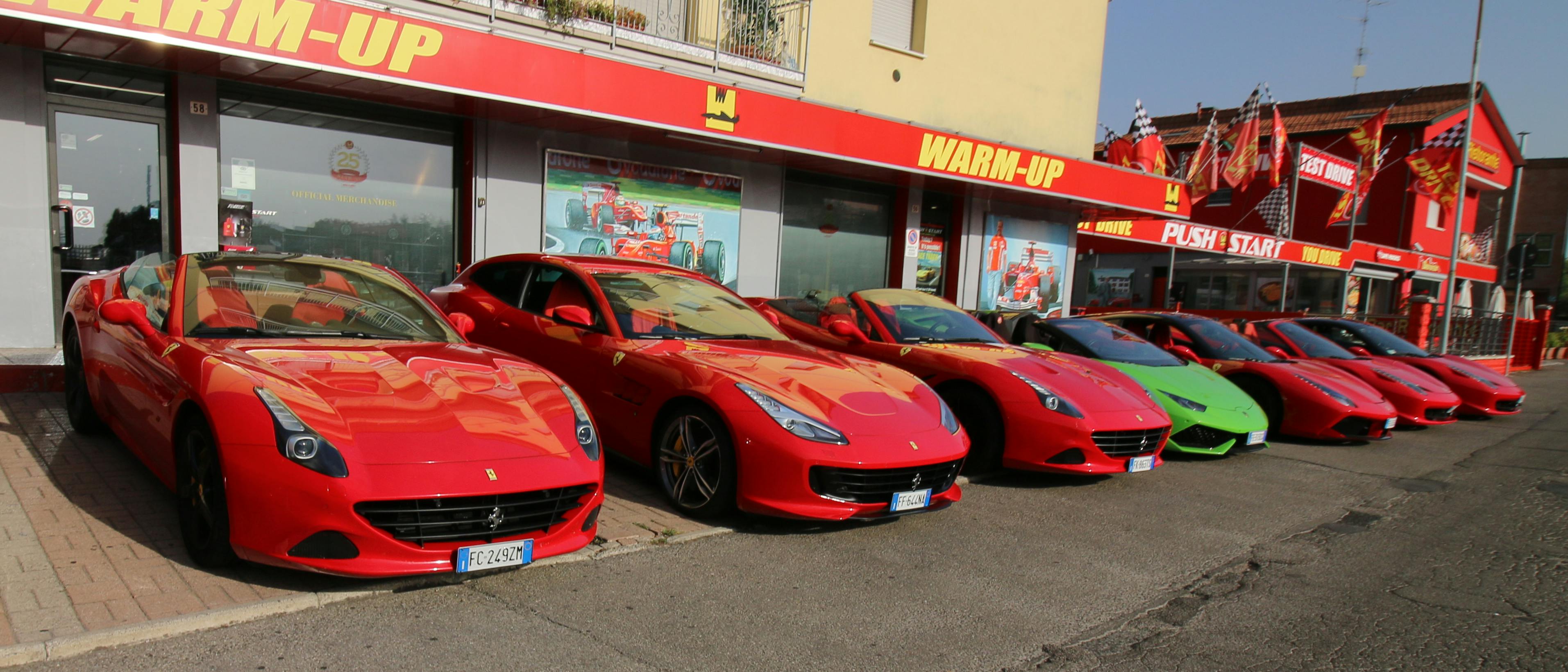 六辆跑车，主要是红色和一辆绿色，停在一家以赛车为主题的装饰的“热身”商店前面。