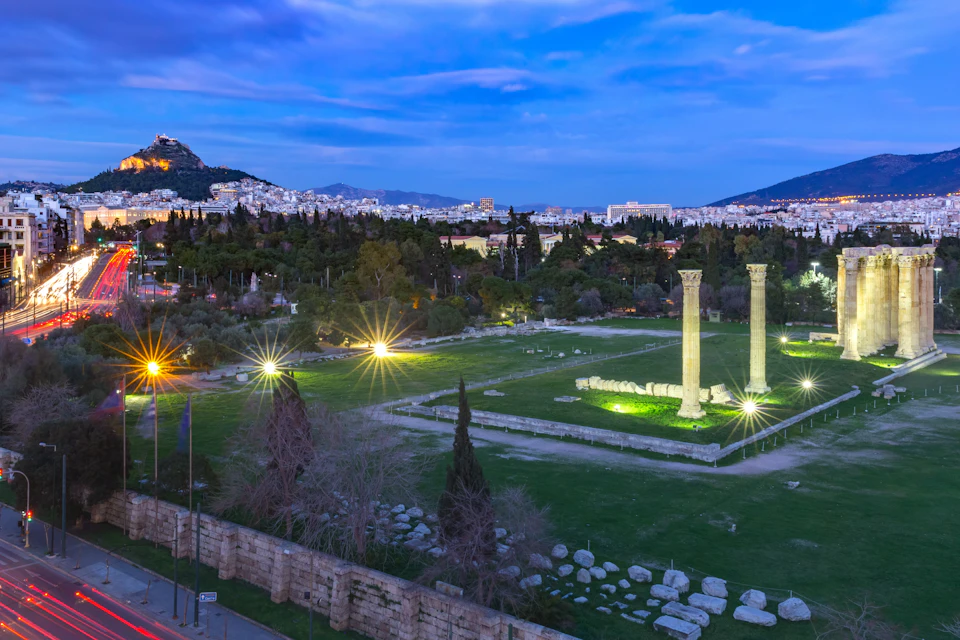 Ancient ruins with two standing columns, illuminated by lights, surrounded by greenery with a city and hills in the background at dusk.