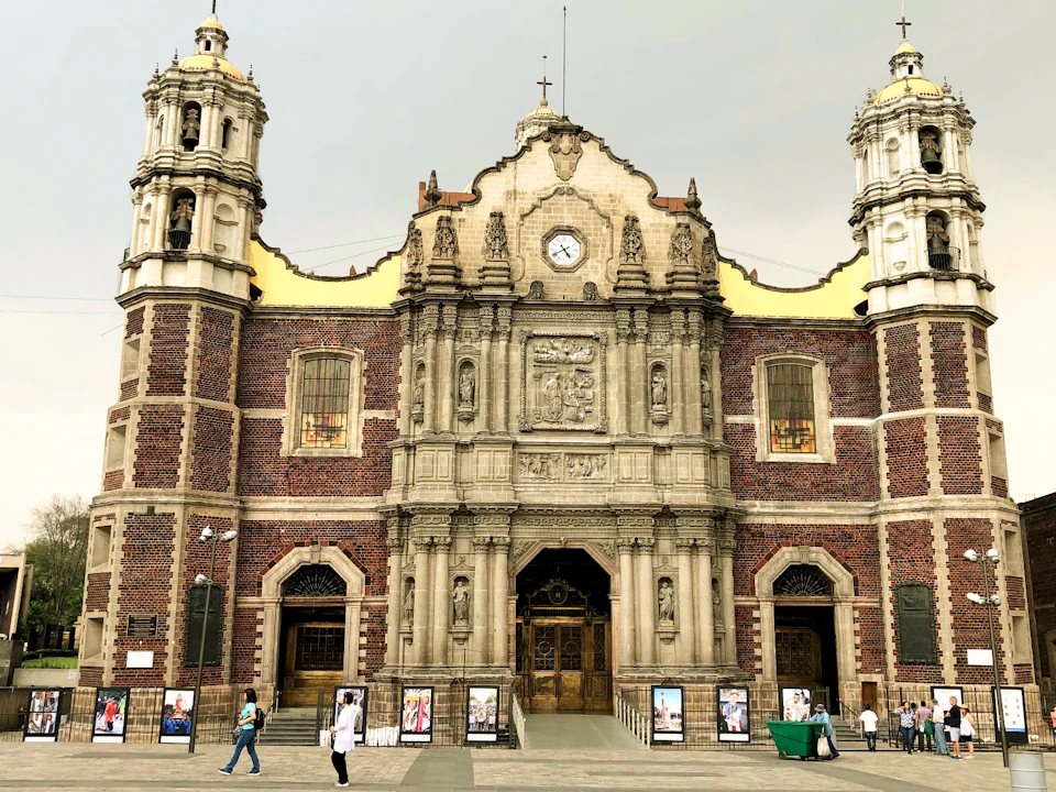 Historic stone church with two towers, clock, ornate central facade, red brick walls; people and posters in front.