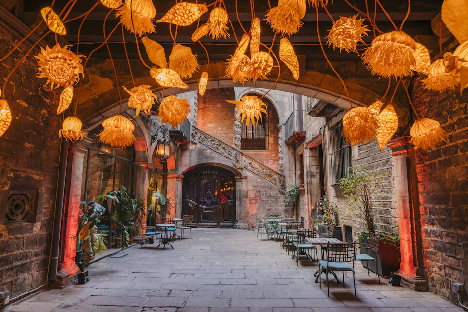 A cozy courtyard with hanging flower-shaped lights, tables, chairs, plants, and an arched doorway under a stone structure.