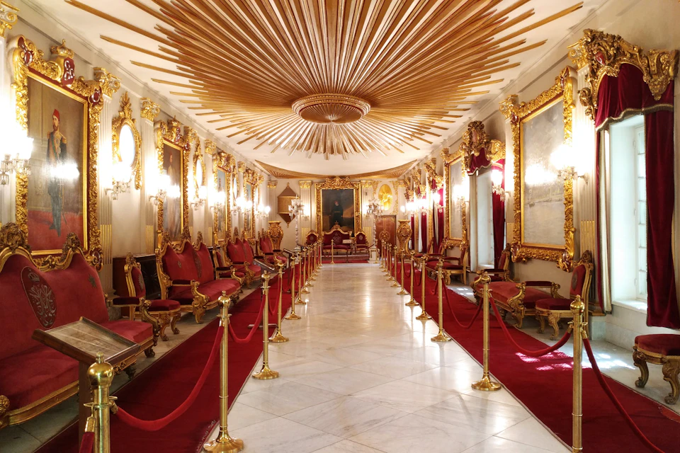 Ornate hall with a red carpet, gold-framed mirrors, chandeliers, red velvet chairs, golden stanchions, and an elaborate ceiling design.