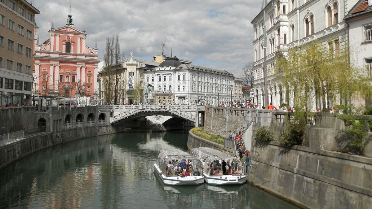 Řeka se dvěma turistickými loděmi a mostem v evropském městě, obklopená historickými budovami pod zataženou oblohou.