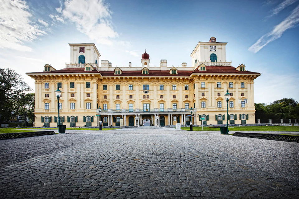 Large, ornate yellow building with tower structures, cobblestone courtyard, lampposts, and blue sky.