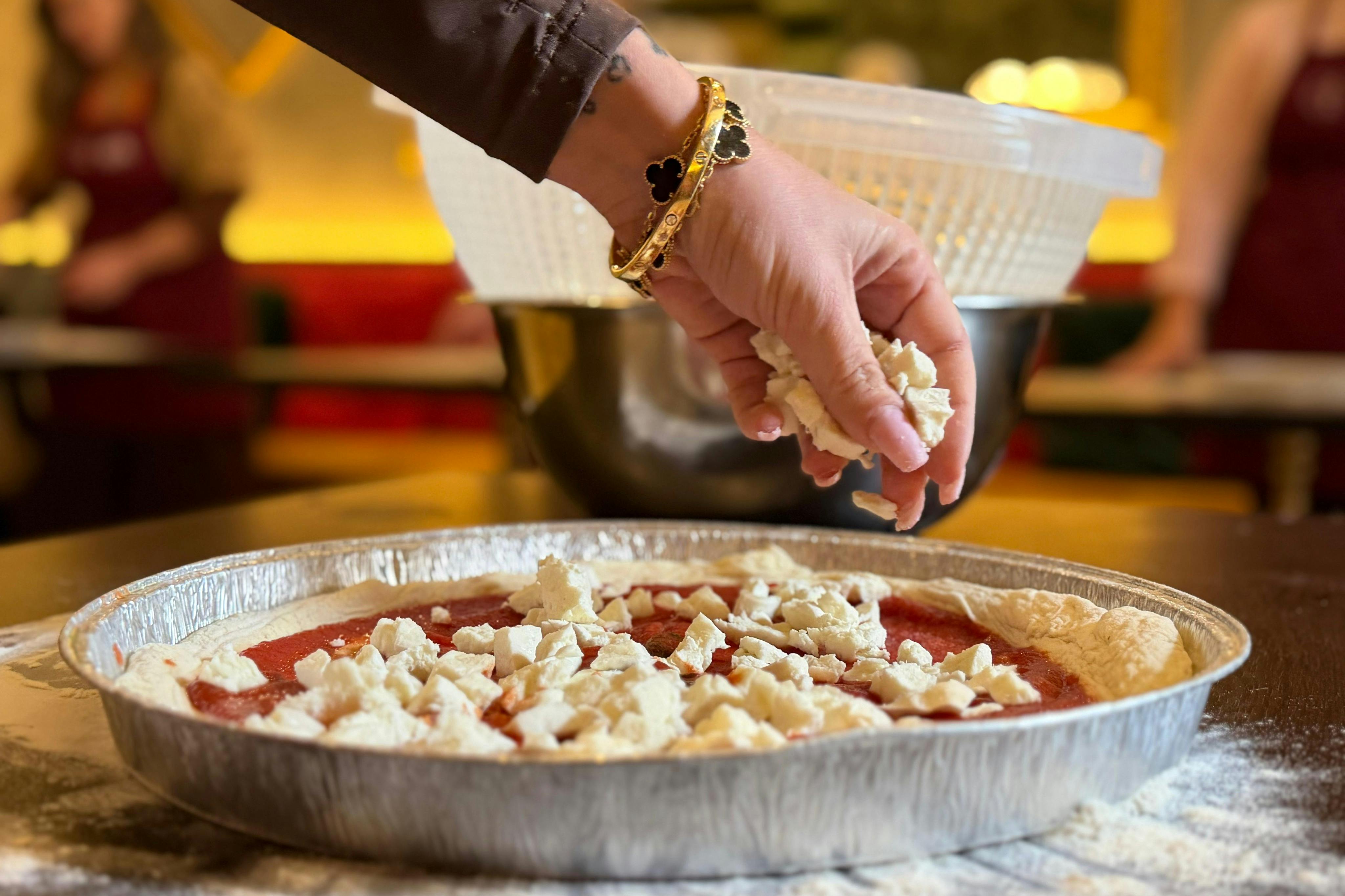 A hand sprinkling cheese onto a pizza in a foil pan, with mixing bowls in the background.
