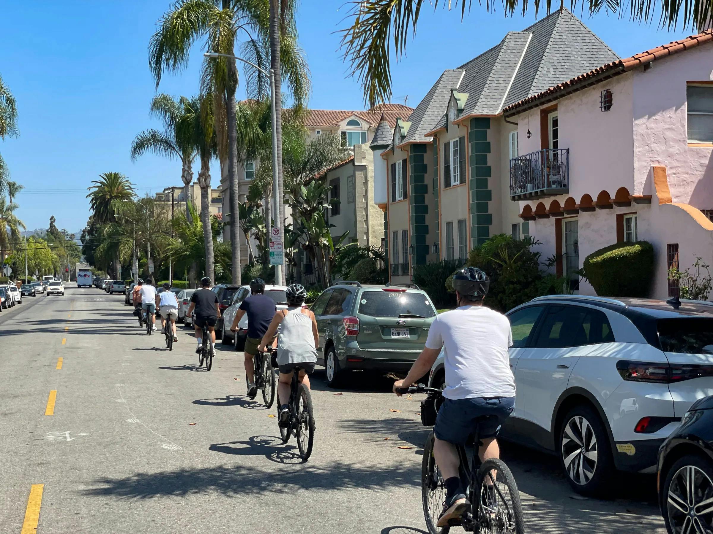 People ride bicycles along a palm tree-lined residential street with parked cars and houses on a sunny day.