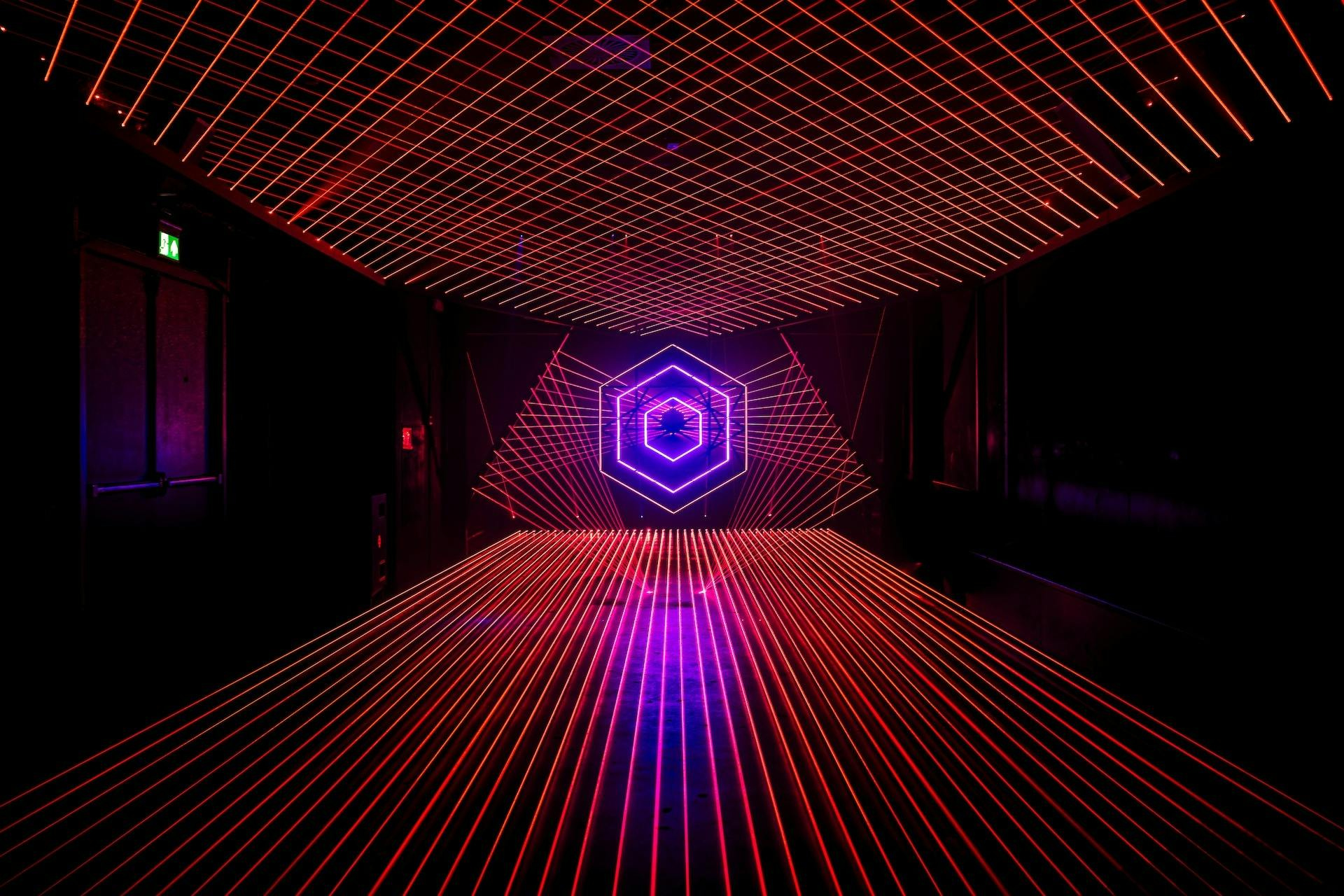 A room with glowing red and pink laser grid lines on the walls, ceiling, and floor, centered around a hexagonal neon pattern.