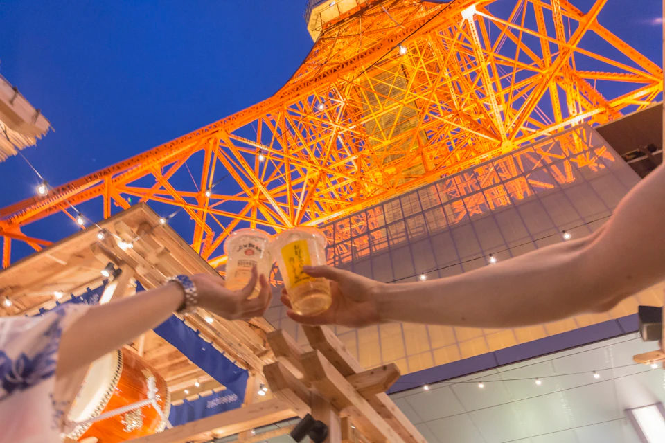 Two people clinking plastic cups of drinks with the illuminated steel structure of Tokyo Tower in the background.