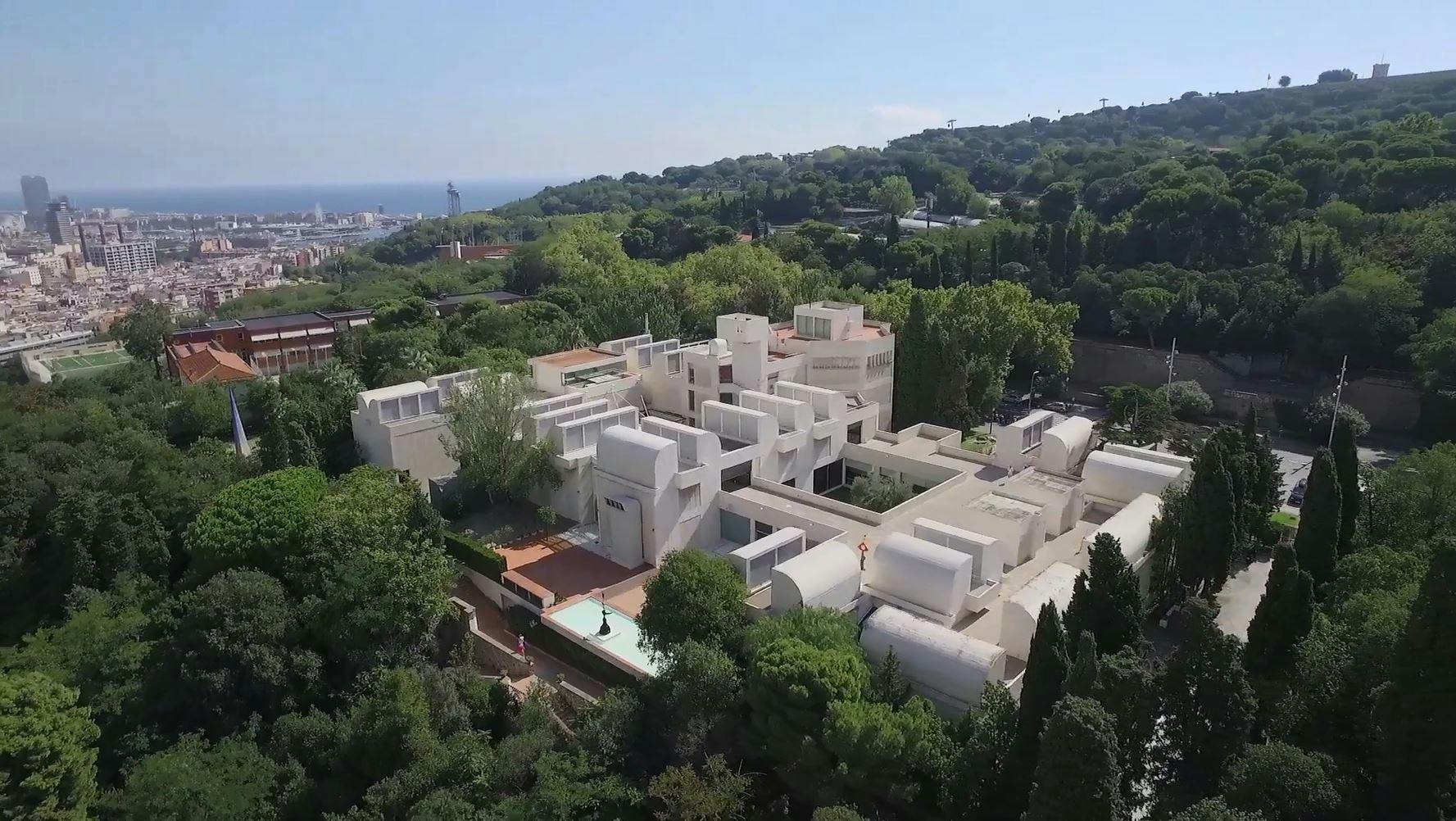 Vit modern byggnad omgiven av grönska, med ett kuperat landskap och havet synligt i bakgrunden.