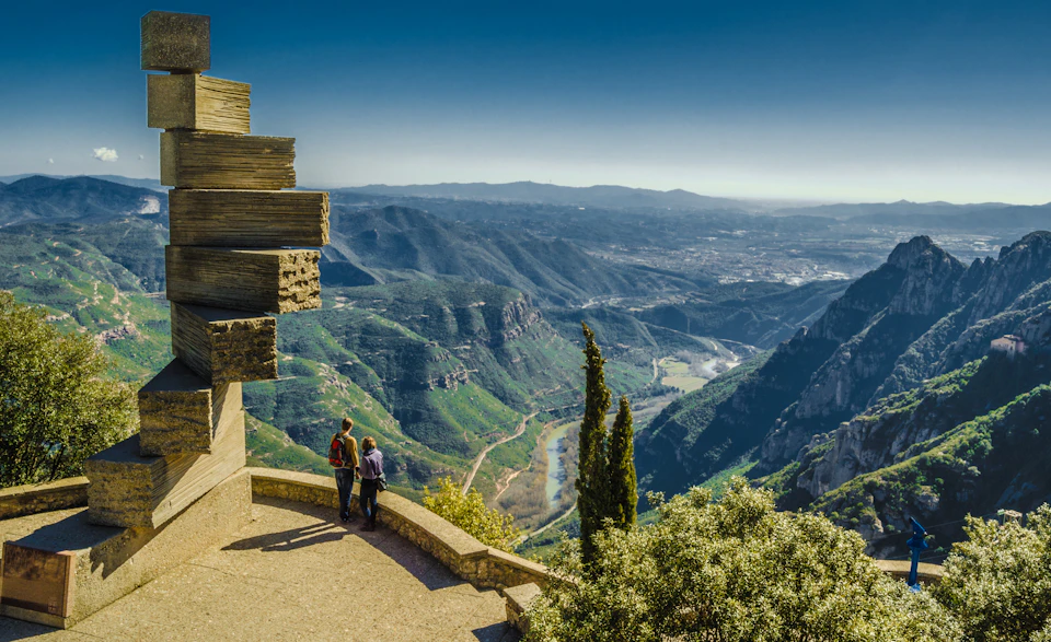 Two people stand on a balcony overlooking a vast valley with mountains, a river, and distant landscape, under a clear blue sky.