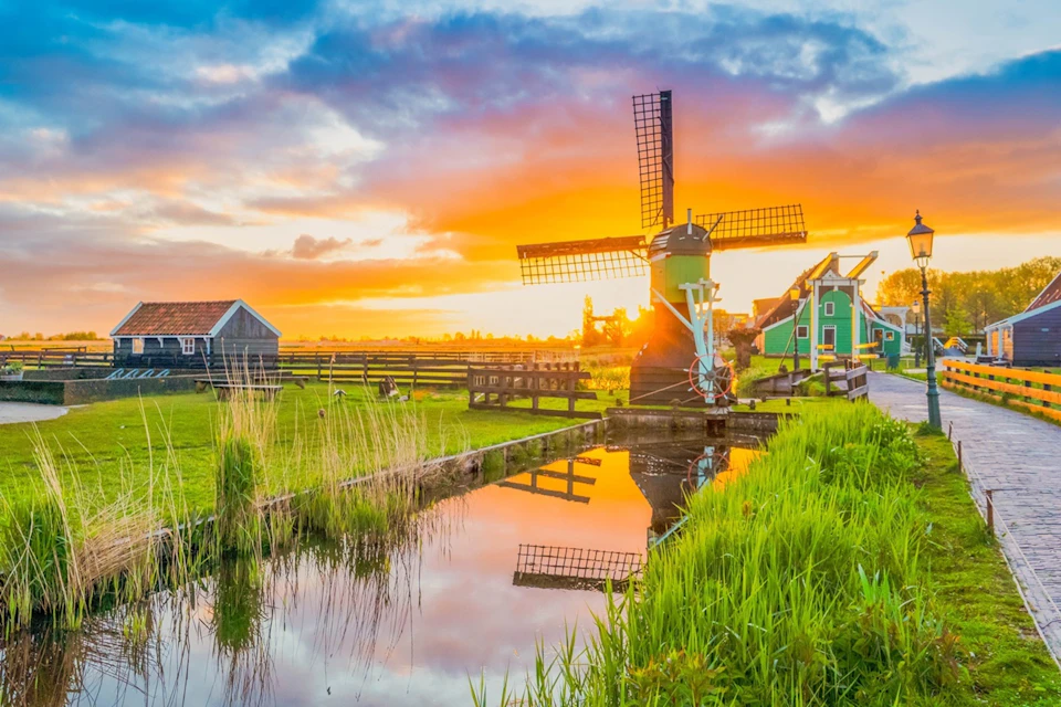 A picturesque sunset over a rural scene with a reflective waterway, windmill, green fields, and quaint houses.
