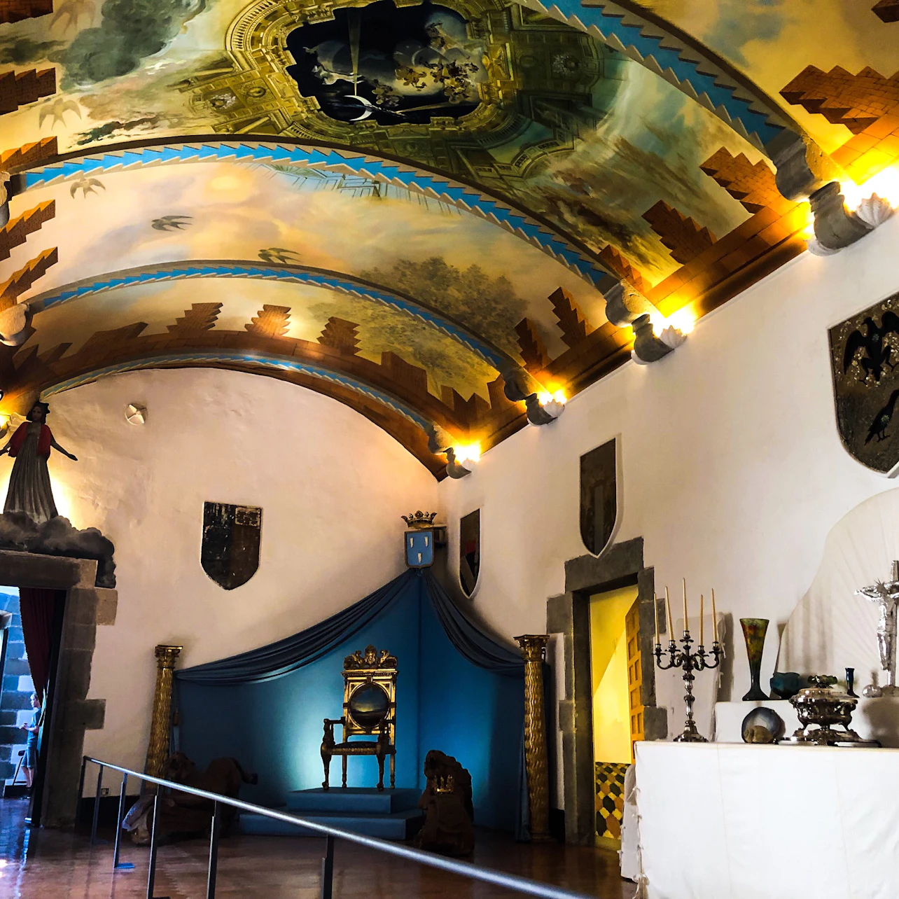 A room with a painted arched ceiling, ornate throne, wall shields, candle holders, and a draped table.