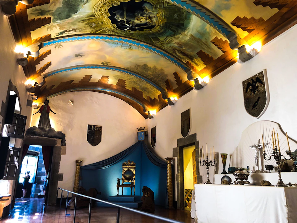 A room with a painted arched ceiling, ornate throne, wall shields, candle holders, and a draped table.