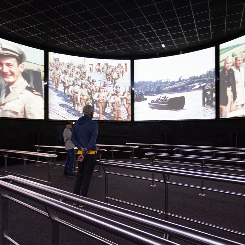 Due persone in piedi davanti a quattro grandi schermi che visualizzano filmati militari storici in una stanza buia con ringhiere di metallo.