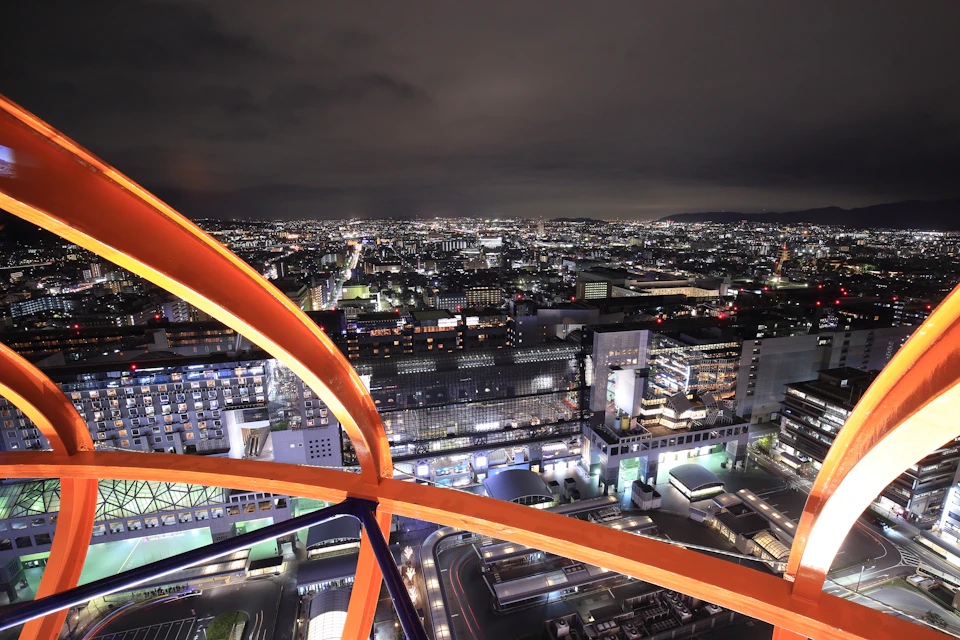 Nighttime cityscape view with illuminated buildings and streets, seen from above through orange metal structures.