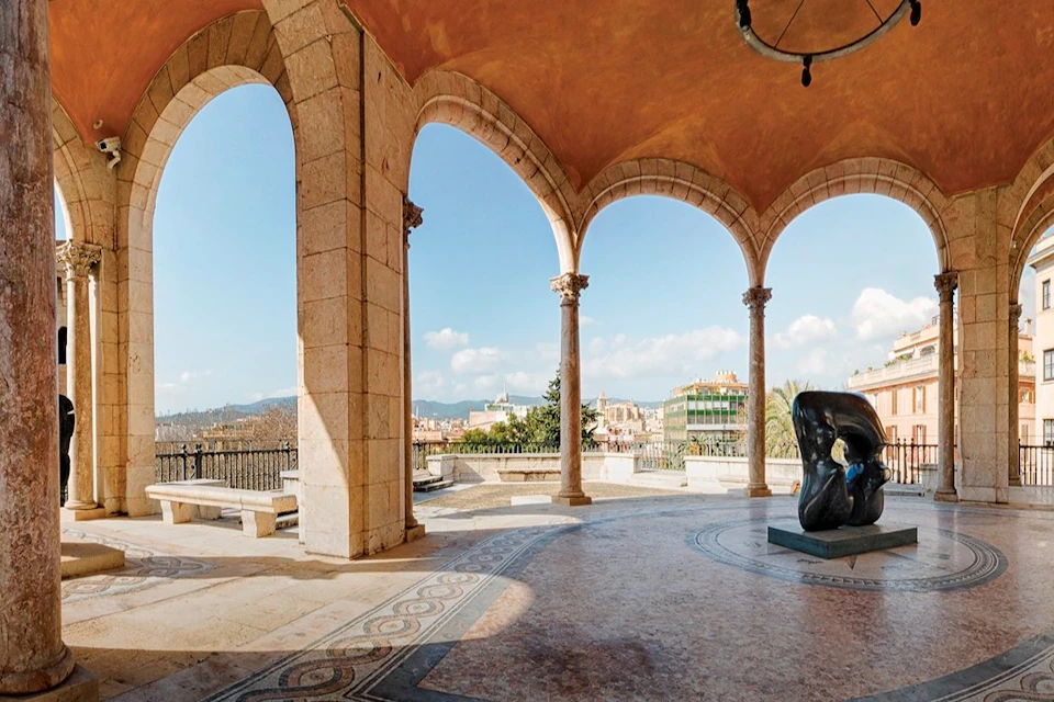 An open colonnaded terrace with stone arches and decorative flooring, overlooking a cityscape and sculpture on a sunny day.