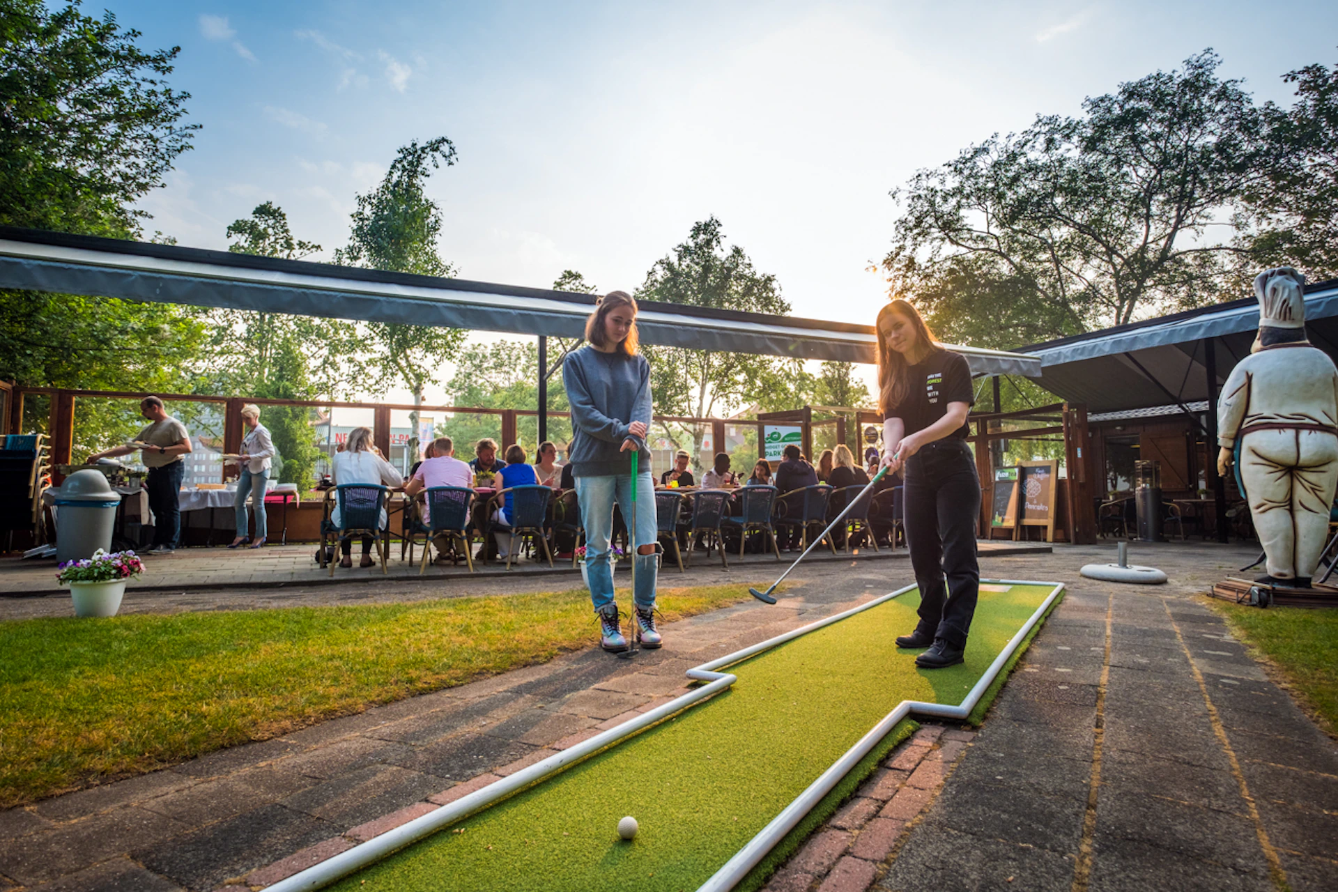 Two people playing mini-golf outdoors, with another group seated at tables under an awning in the background.
