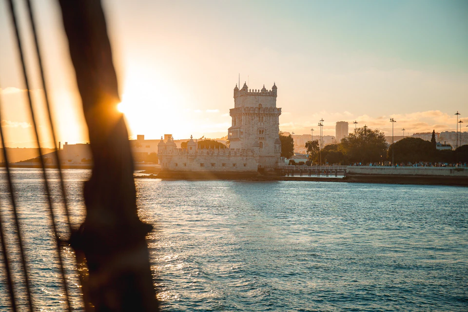 A historic stone tower by the water at sunset, surrounded by trees and buildings in the distance.