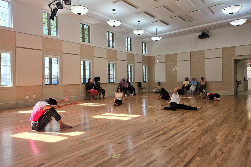 A spacious room with people seated and performing dance or stretching poses on a wooden floor, with natural light through windows.