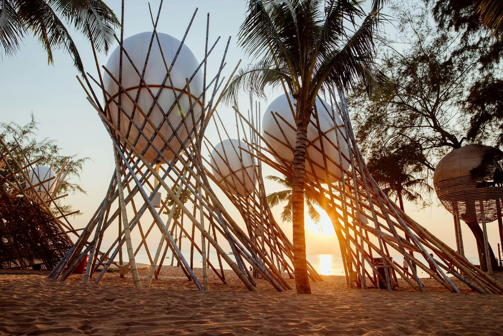 Large white spherical structures supported by bamboo frames on a sandy beach with palm trees at sunset.