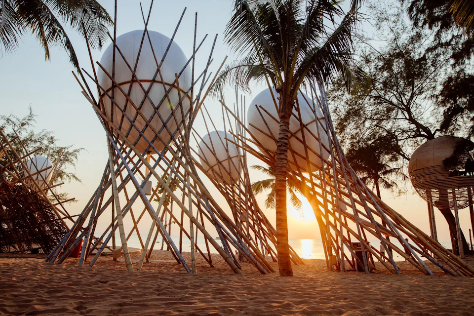 Large bamboo structures with spherical tops on a sandy beach during sunset, surrounded by palm trees.