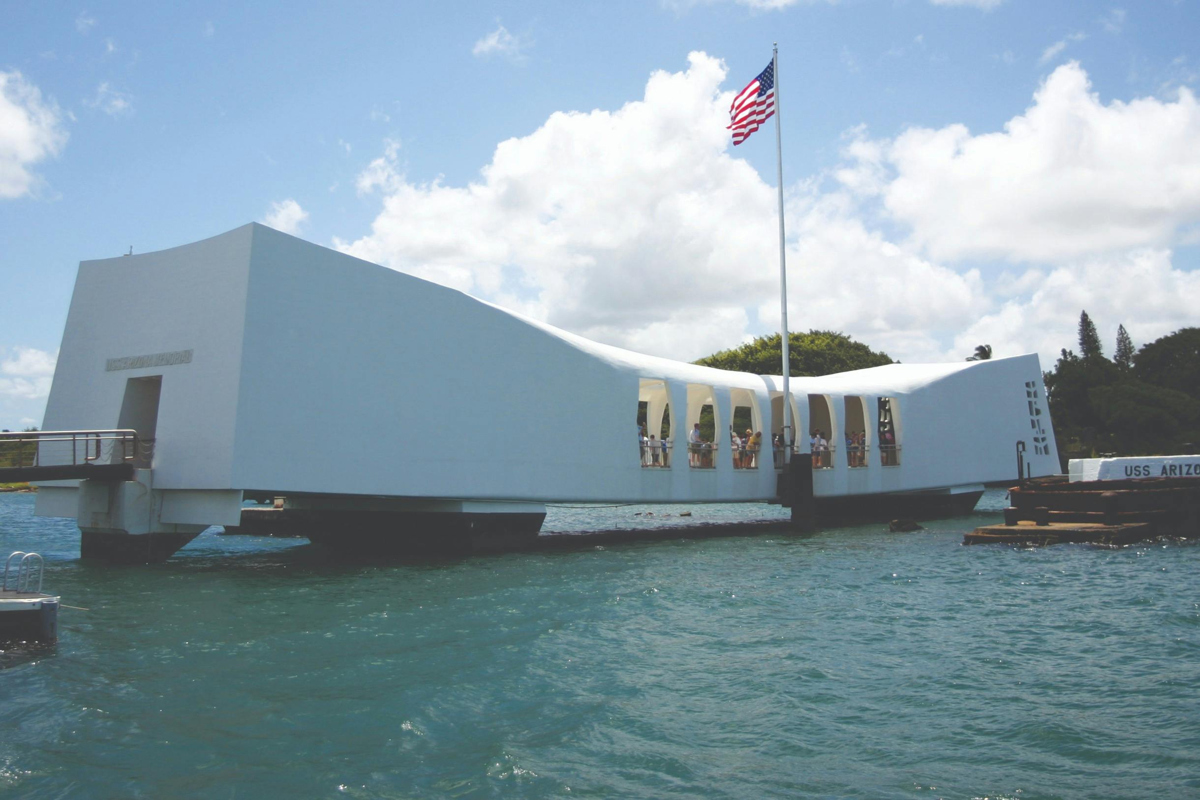 A white memorial structure over water with an American flag atop a pole, under a partly cloudy sky.