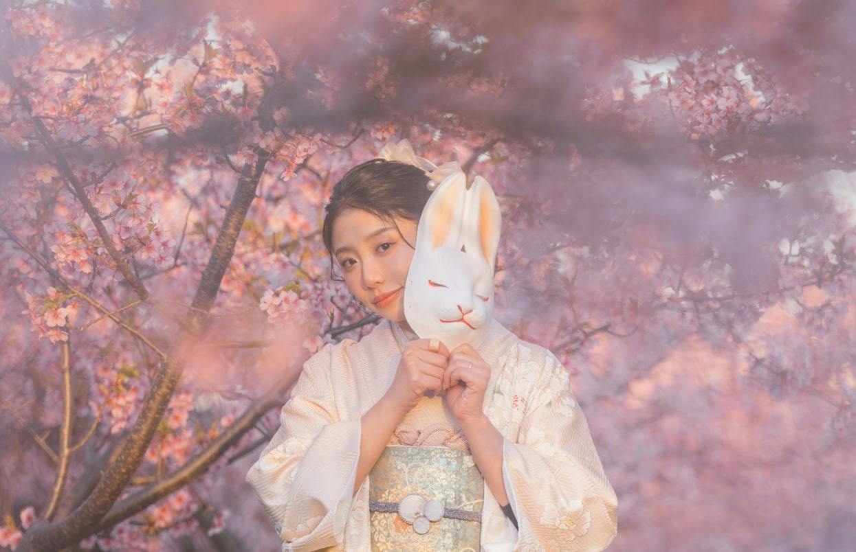 A person in a traditional outfit holding a rabbit-shaped mask amidst blooming cherry blossoms.