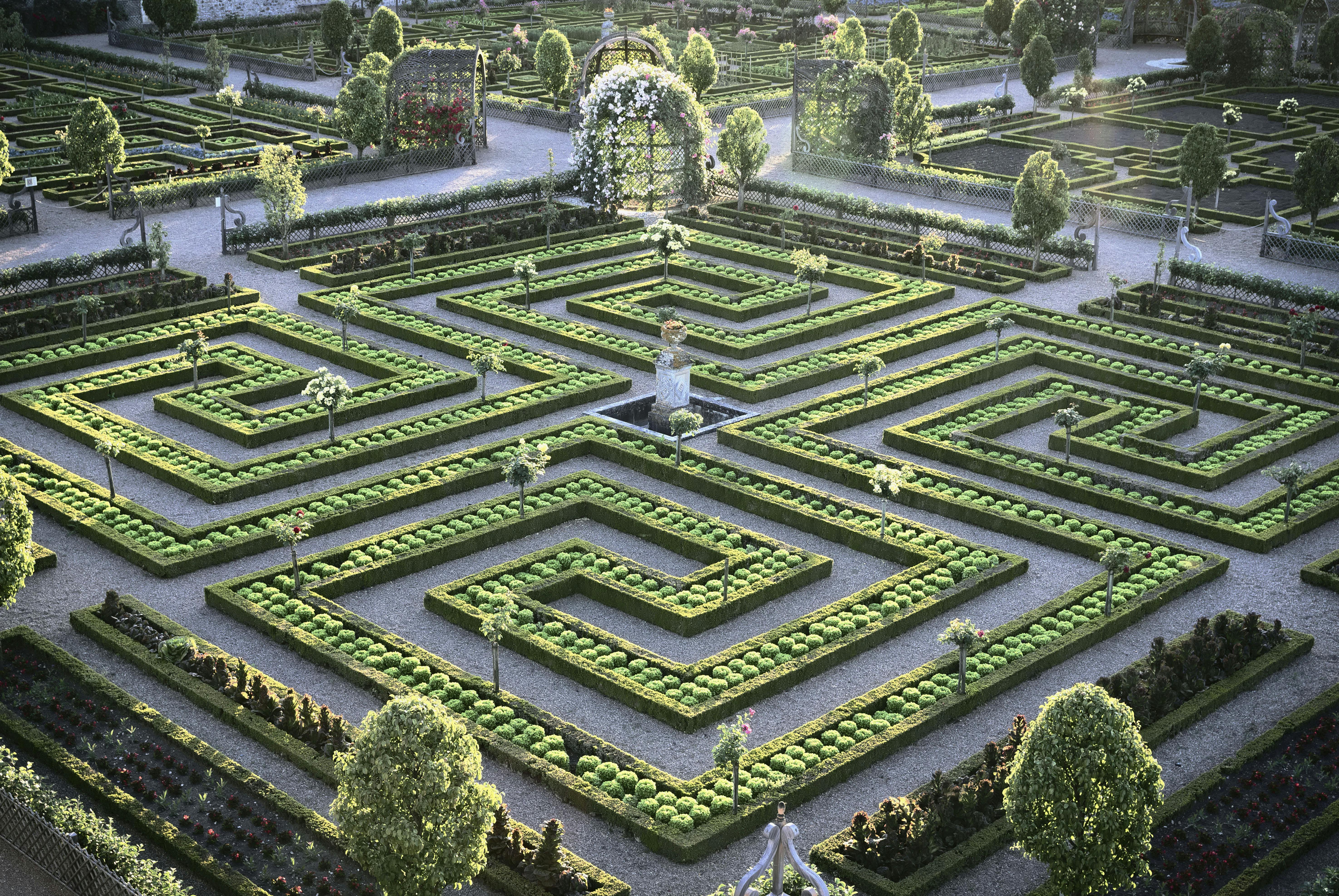 Un labirinto da giardino ornato con siepi tagliate, alberi topiati, aiuole e una statua centrale sotto un cielo soleggiato.