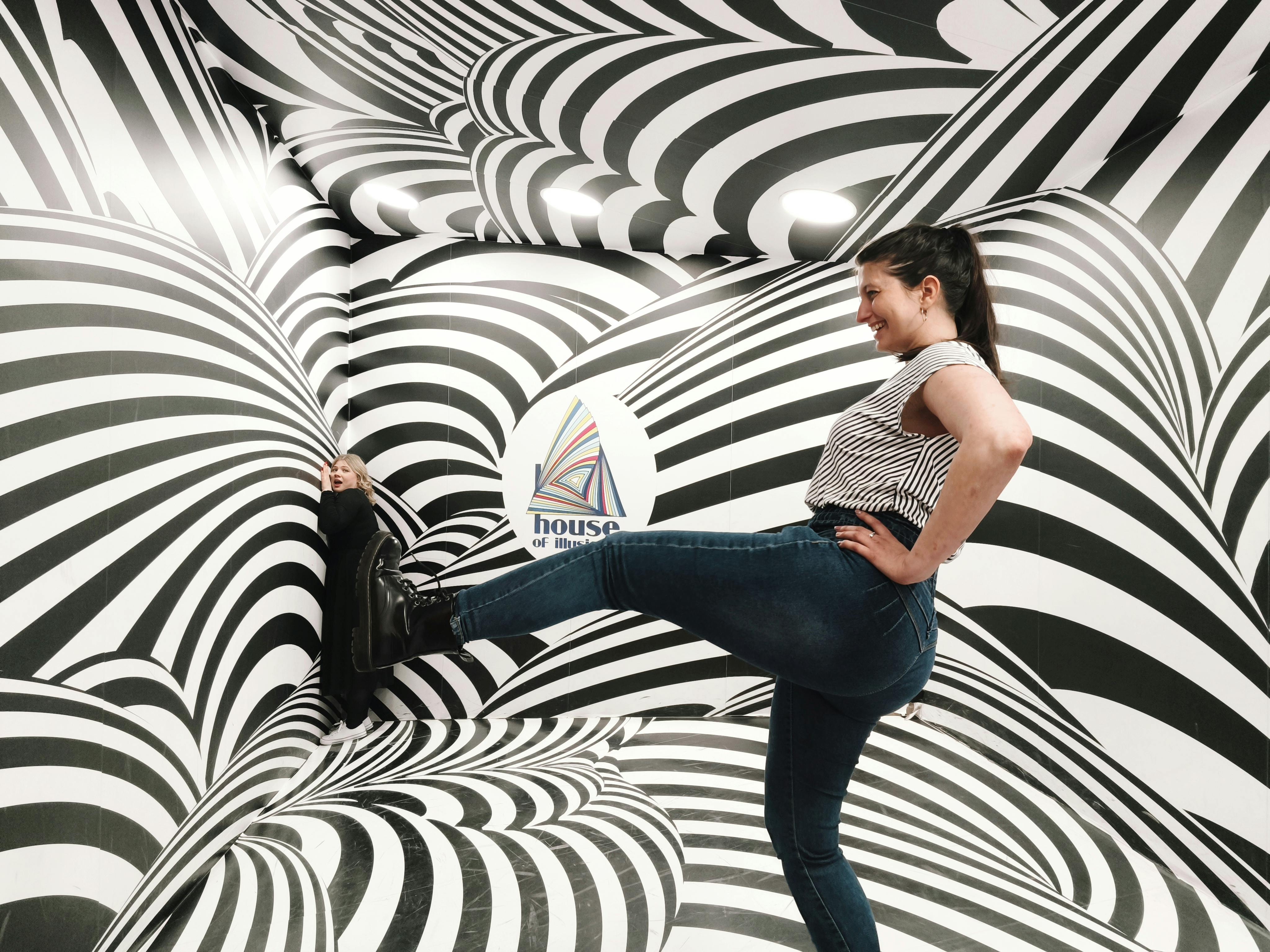 Une femme pose de manière ludique dans une pièce dont les murs et le sol sont ornés de motifs tourbillonnants noirs et blancs.