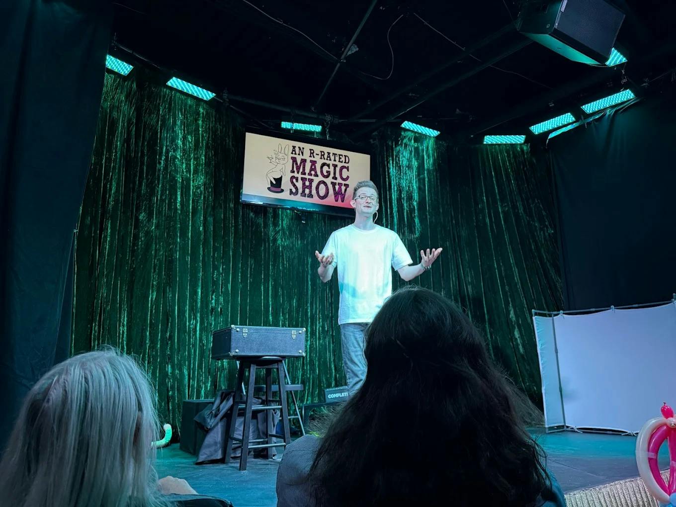 Un mago con camisa blanca actúa en el escenario con un telón verde de fondo y un cartel que dice "An R-Rated Magic Show".
