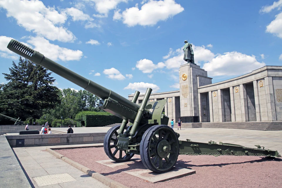 Large artillery gun on display in front of a monument featuring a statue and plinth, with several people nearby under a blue sky.