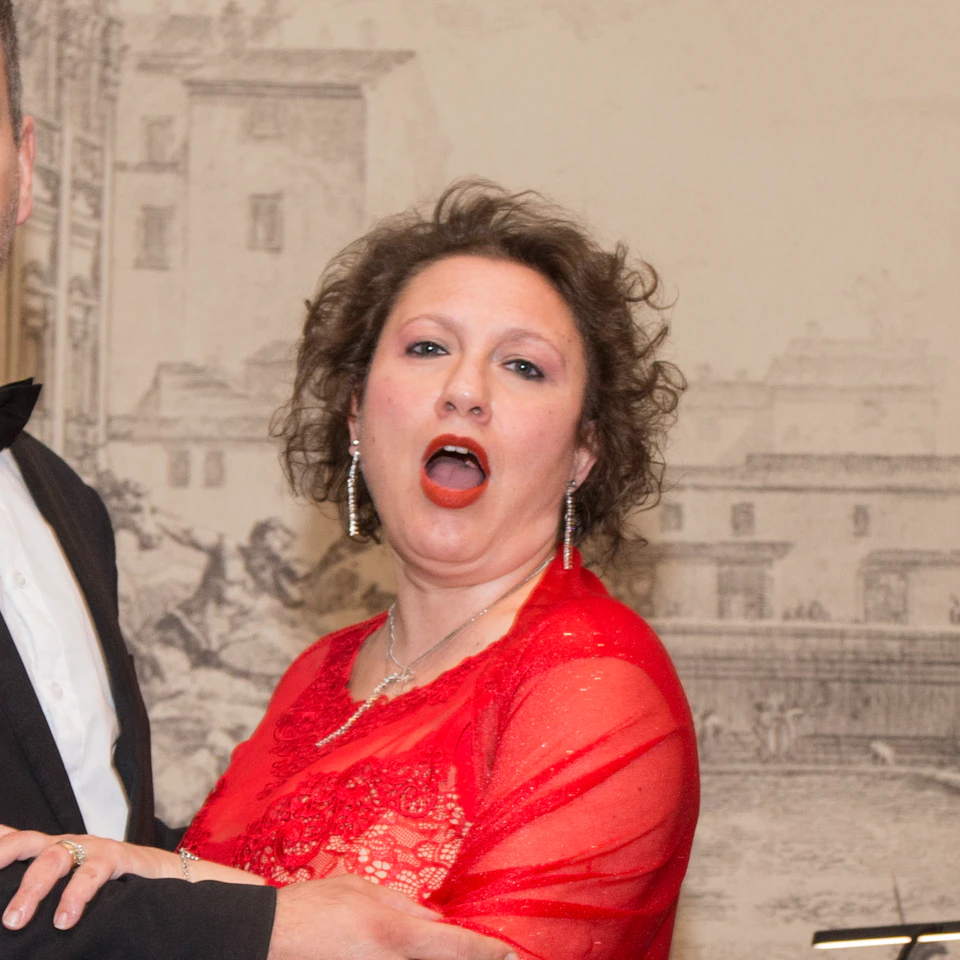A man in a tuxedo and a woman in a red dress perform in front of a classical backdrop, with musicians playing in the background.