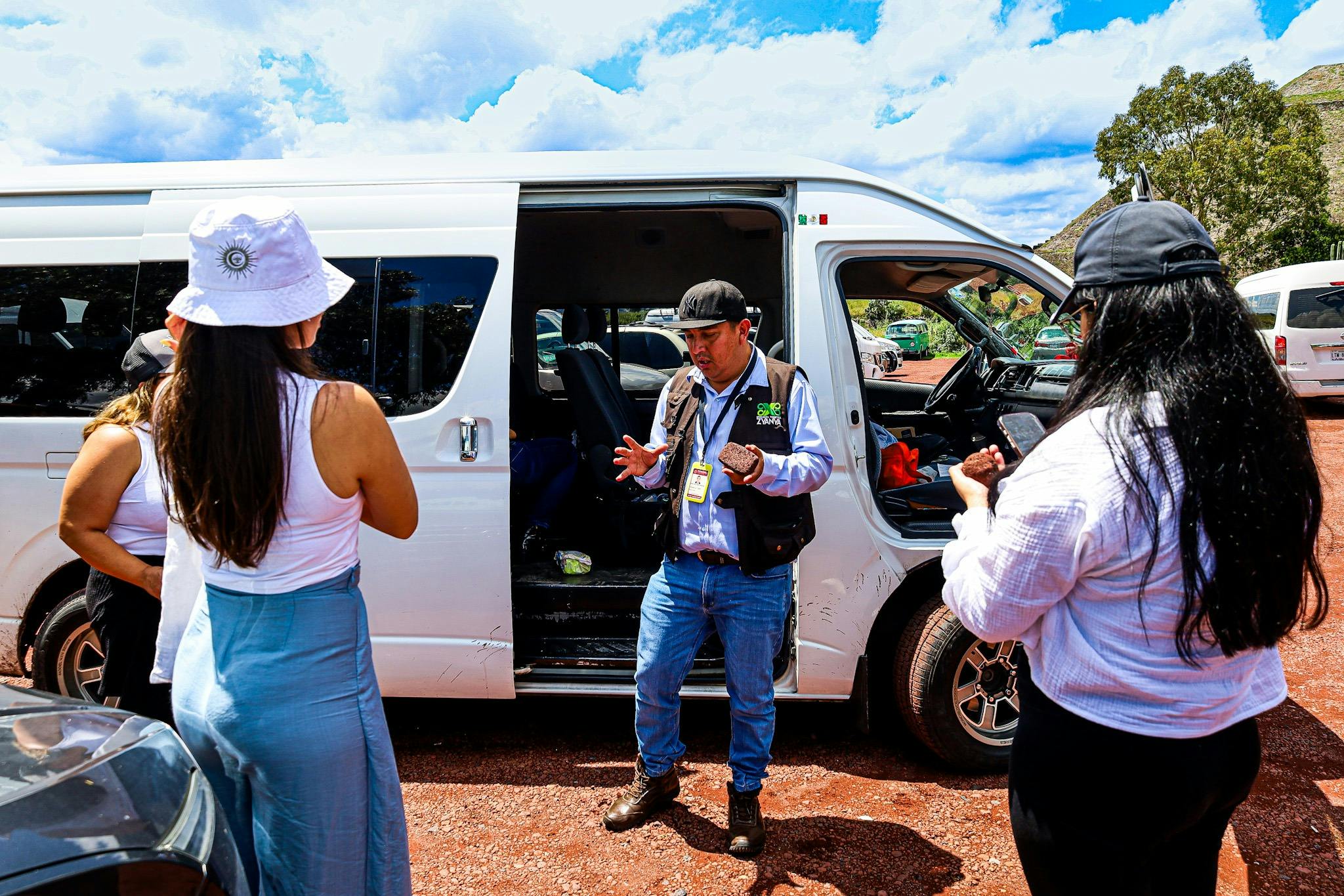 A guide speaking to a group near a white van with its sliding door open, under a sunny sky with clouds.