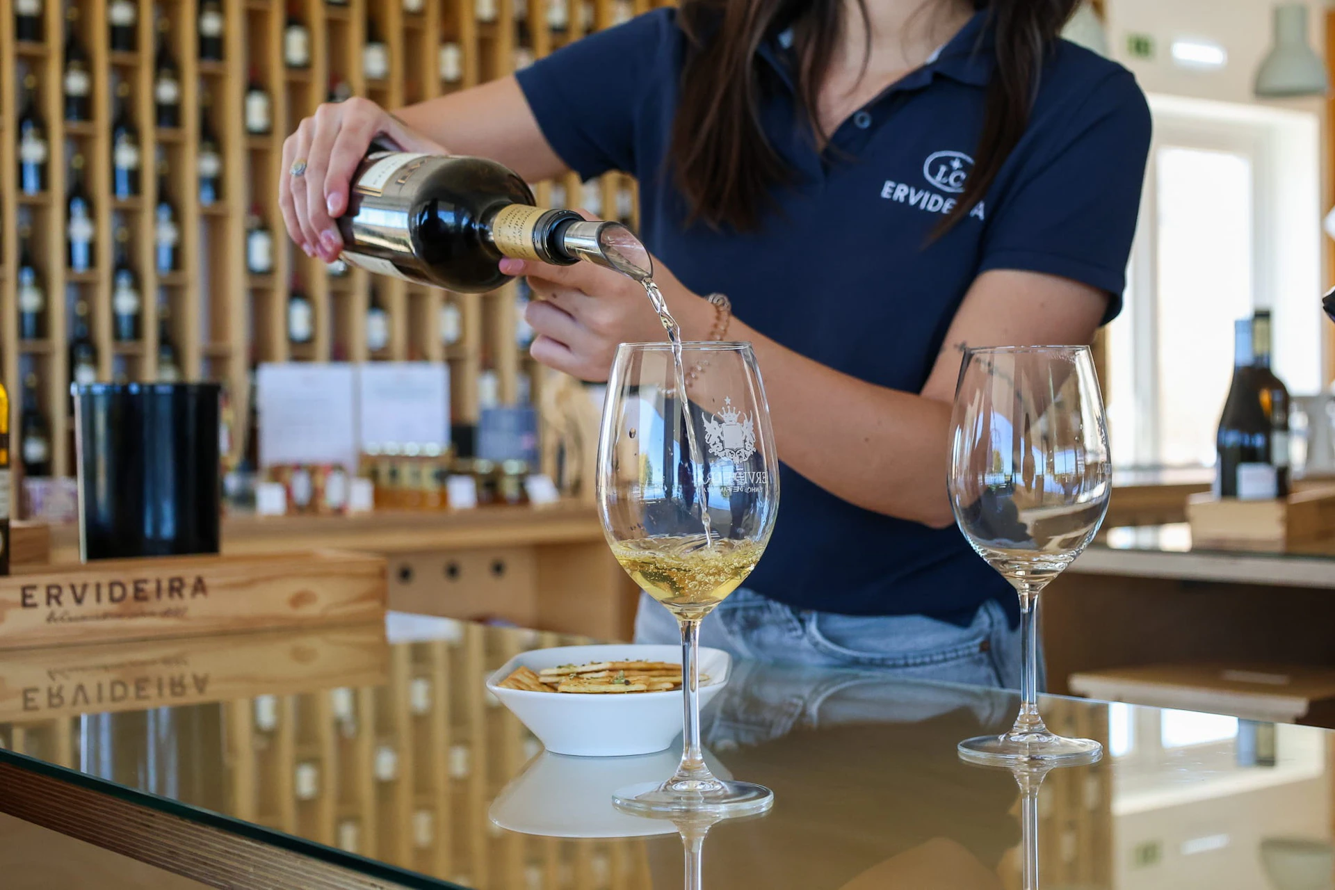 A person in a blue polo shirt pours white wine into a glass on a counter with crackers and empty glasses.